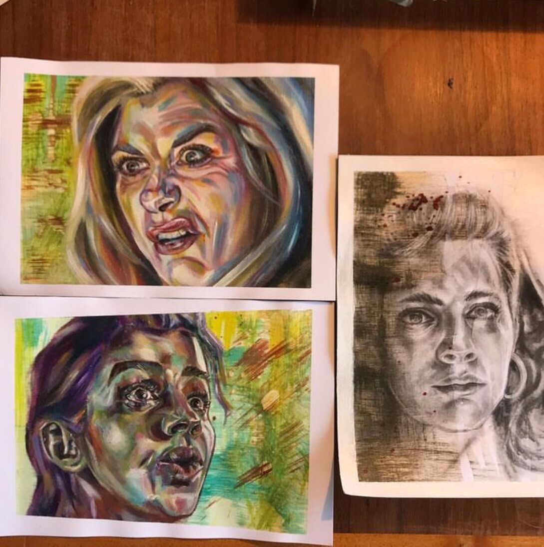3 paintings on paper on a desk. Two are colorful interpretations of female characters from Dr Who. One is a graphite and acrylics portrait of a female character from Deadly class
