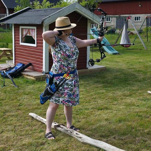 En bågskytt i klänning och solhatt. I bakgrunden, en lekstuga, gungställning, rutschkana och en lagugård.