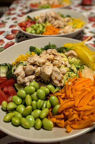 A plate in the foreground filled with vegetables like carrot, edamame, cucumber, bell pepper, corn, broccoli, lettuce and sushigari, topped with stir-fried chicken. Two more plates are out of focus in the background.