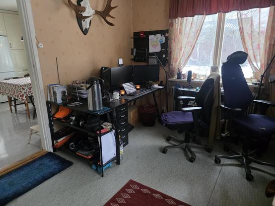 A desk in a corner, not too cluttered, with three screens. A window in the background hints at snow outside, and two small candles are lit on the desk on either side of the tablet in front of the screens. The billboard in the corner has few items, hinting at a not too busy schedule at the moment. It is a restful winter-setup.