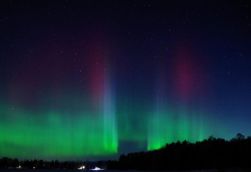 A colourful image of aurora borealis above a silhouette of the forest. At the low part some strong green , a bit higher up in the starry sky the red aurora appears really strong red and at the center it almost looks as if there is a portal with pillars on each side where the pillars to the left is almost white. The sky is filled with stars.