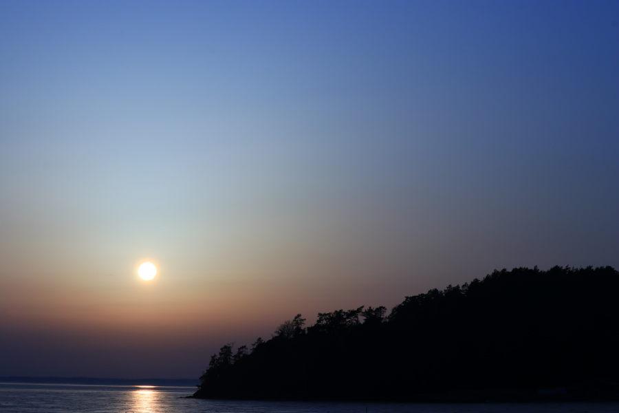 The sun behind some haze aboute an hour before sunset. The upper sky is cold light blue and the lower part is warm orange and dark purple. The sunlight is reflected on the surface of the sea, and due to the haze it looks more like an image of the moon than the sun. To the rigth a headland hill with forest in a dark silhouette.