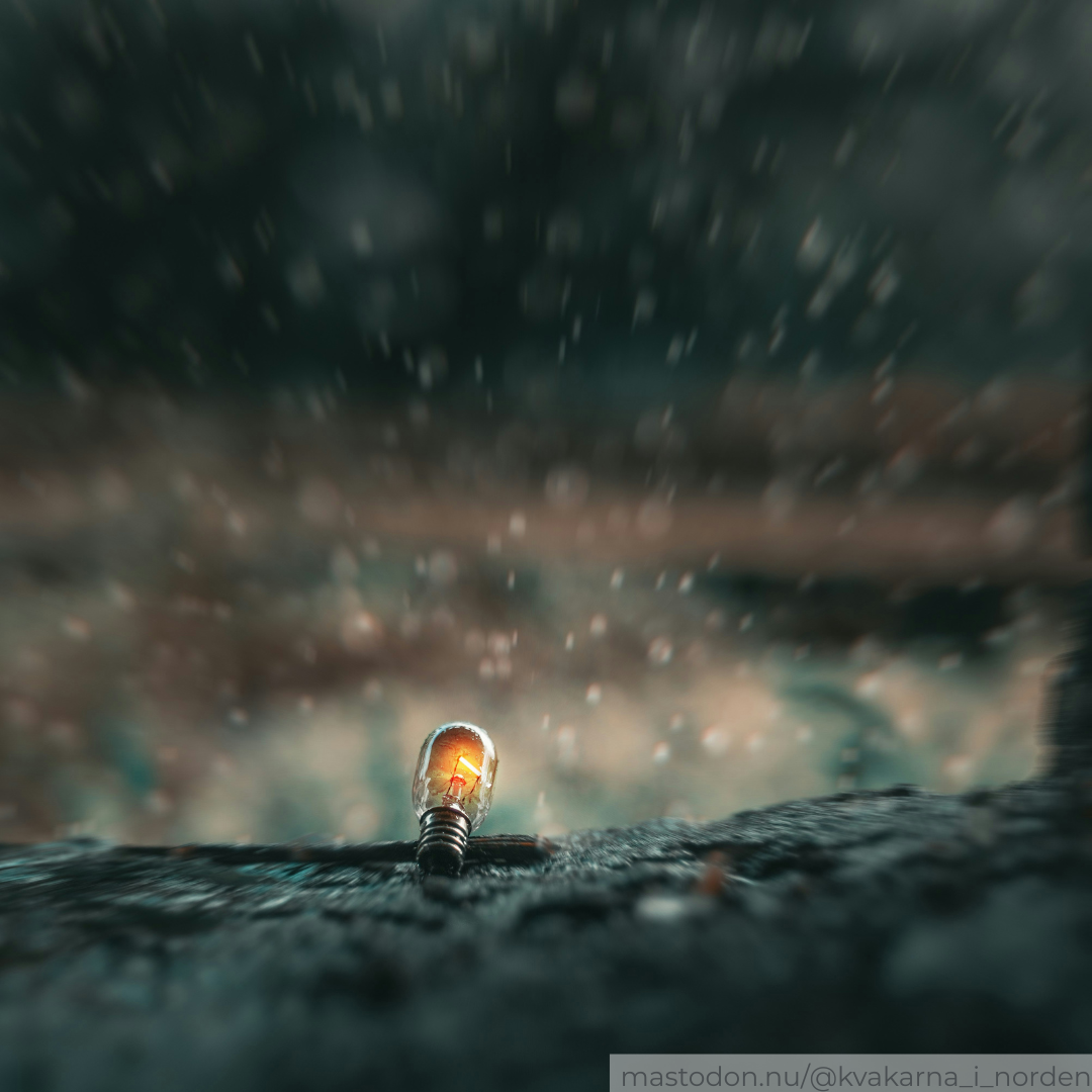 ​A moody, macro photograph of a tiny, incandescent light bulb resting upright on a rough, textured dark surface. The bulb glows with a warm orange light from its internal filament. Heavy rain or water droplets fall all around it, creating a strong bokeh effect with blurred streaks and droplets against a dark, out-of-focus background. A small watermark in the bottom right corner reads "mastodon.nu/@kvakarna_i_norden".