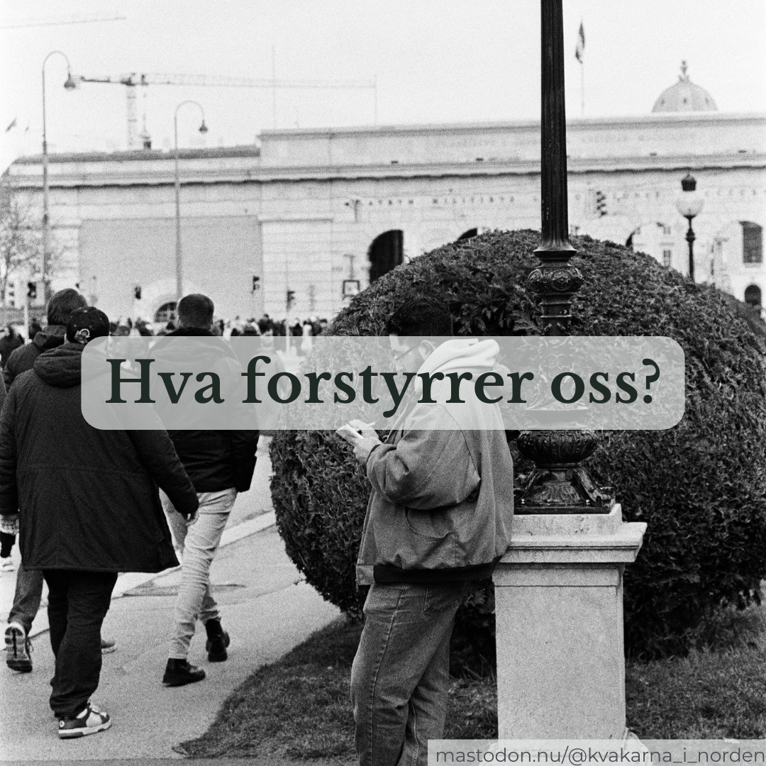 ​A black and white street photograph showing a man standing in profile, leaning against the stone base of a lamppost while focused on his smartphone. In the background, other pedestrians walk past a large, rounded green bush, with grand classical architecture and a construction crane visible further back. A semi-transparent overlay box across the center contains the Norwegian text, "Hva forstyrrer oss?". A small watermark in the bottom right corner reads "mastodon.nu/@kvakarna_i_norden".