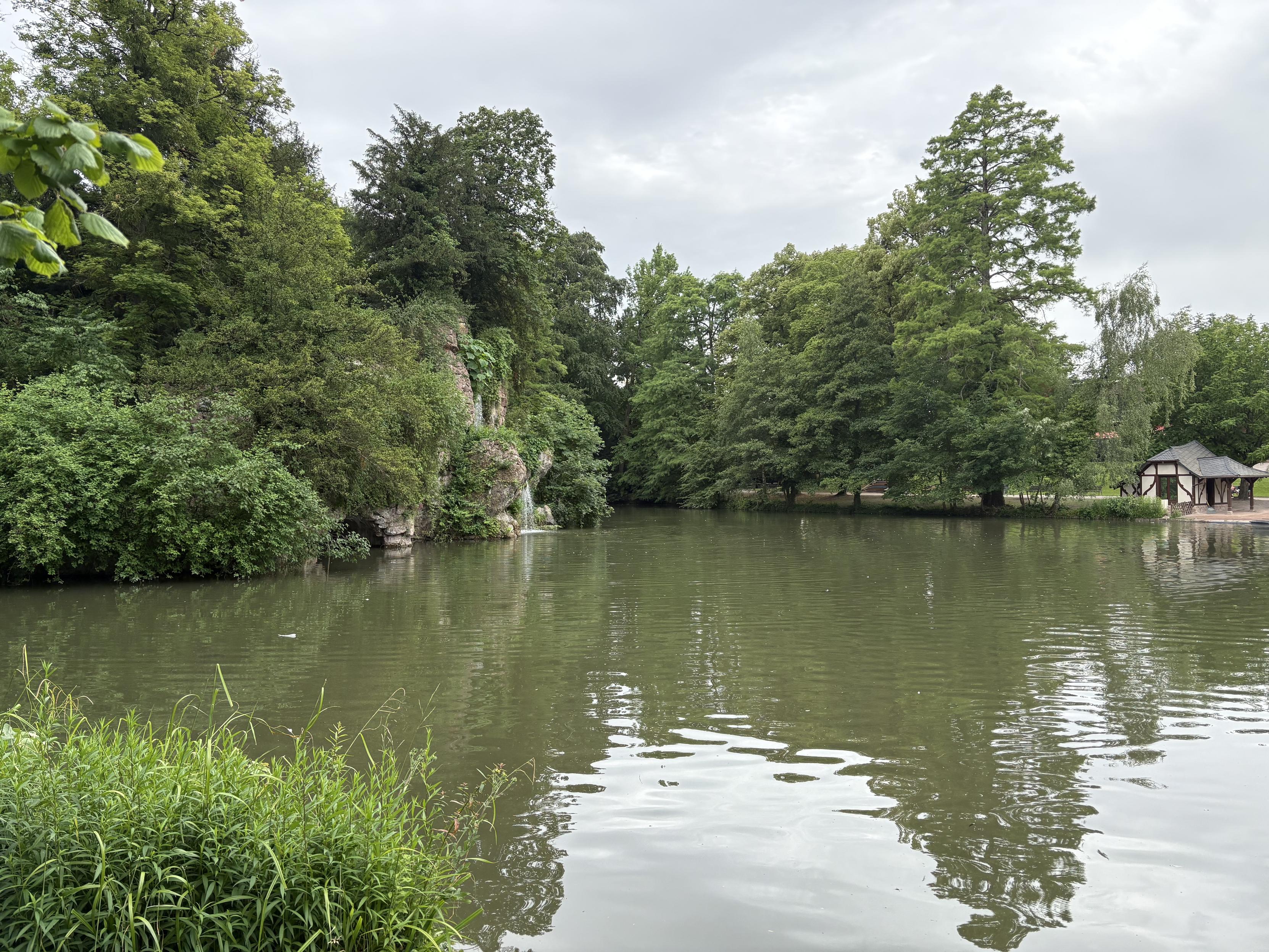 Parque con un lago natural rodeado de árboles. Una cascada cayendo de unas piedras a la izquierda. Una casa de tipo tradicional alsaciano en la orilla a la derecha.