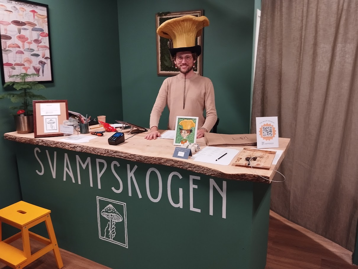En leende butiksinnehavare står stolt bakom sin disk med en svampliknande hatt på huvudet.