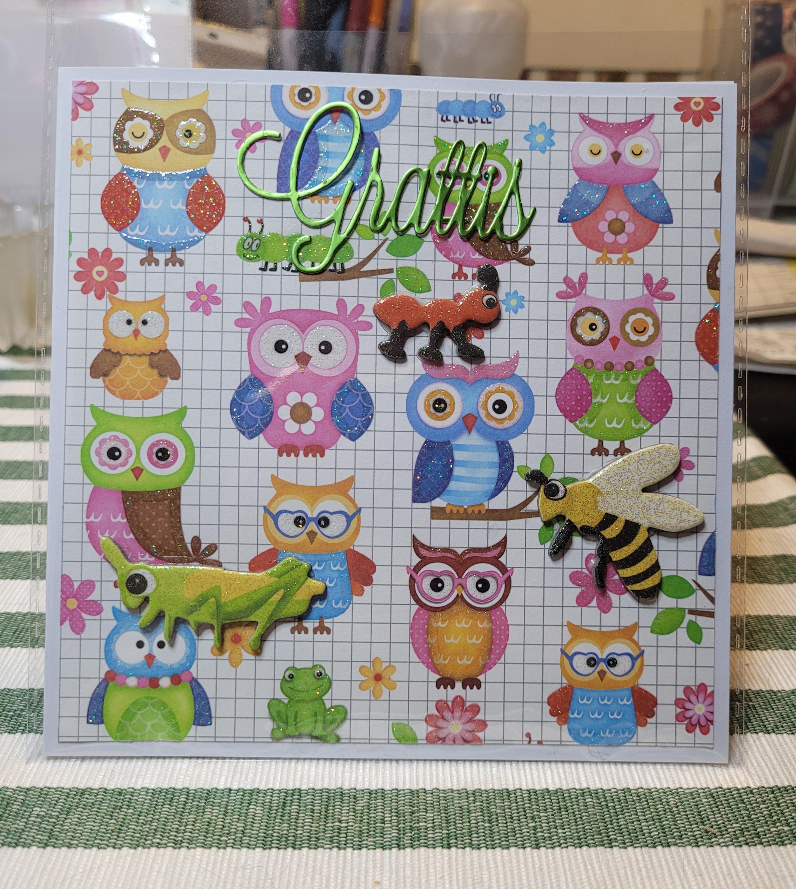 Grattiskort med ugglor i olika färger på vit bakgrund några gröna larver och en groda samt blommor syns också.
En gräshoppa ett bi och en myra i glittrig cardboard sitter och på kortet.
Grattis utstansat  i grönt metallic papper