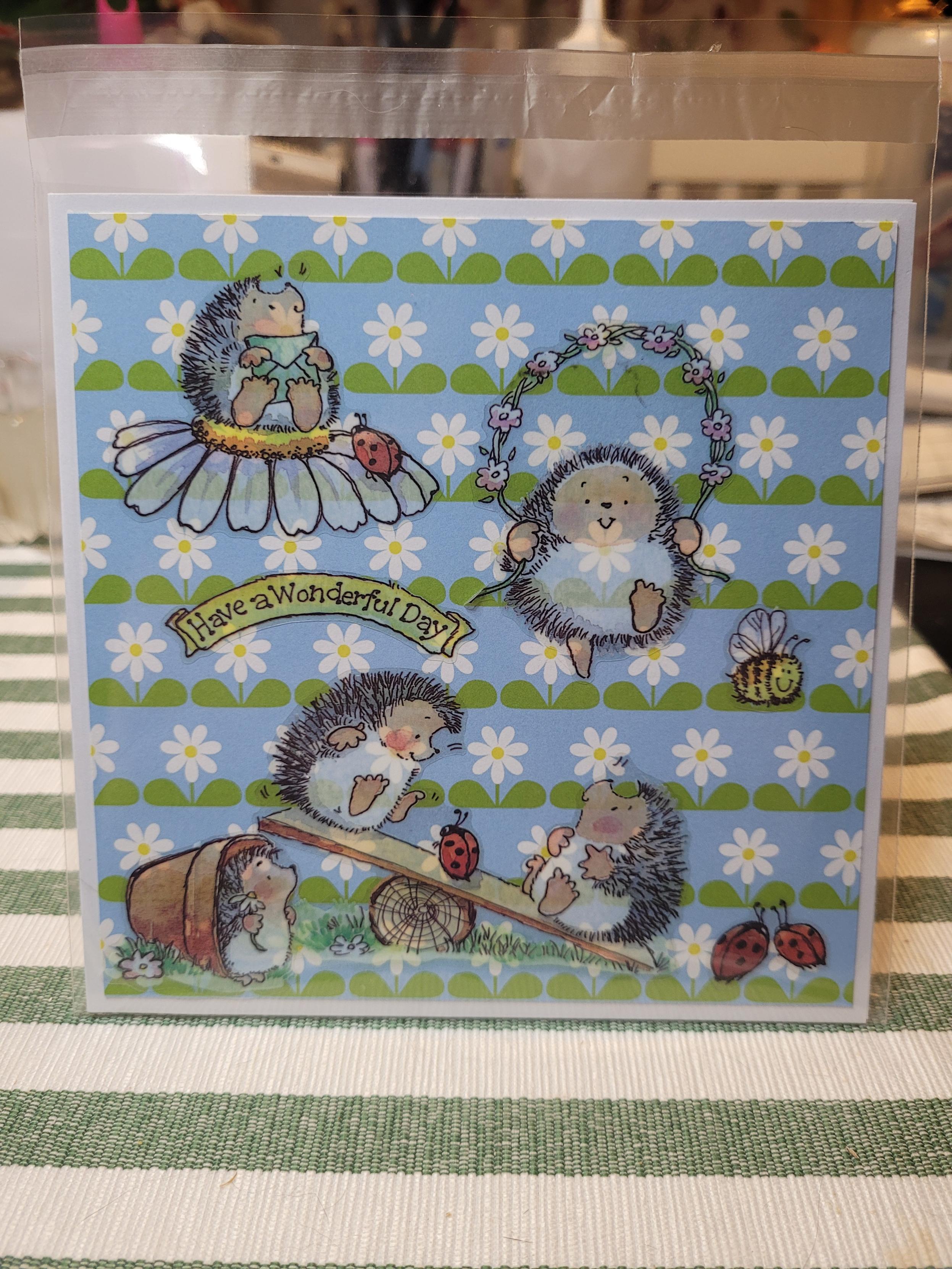 Grattiskort  med igelkottar i olika positioner i en trädgård.
"Have a wonderful day " i gult med svarta bokstäver.
En humla och nyckelpigor syns också.
Bakgrunden är blå botten med vita blommor och gröna blad.