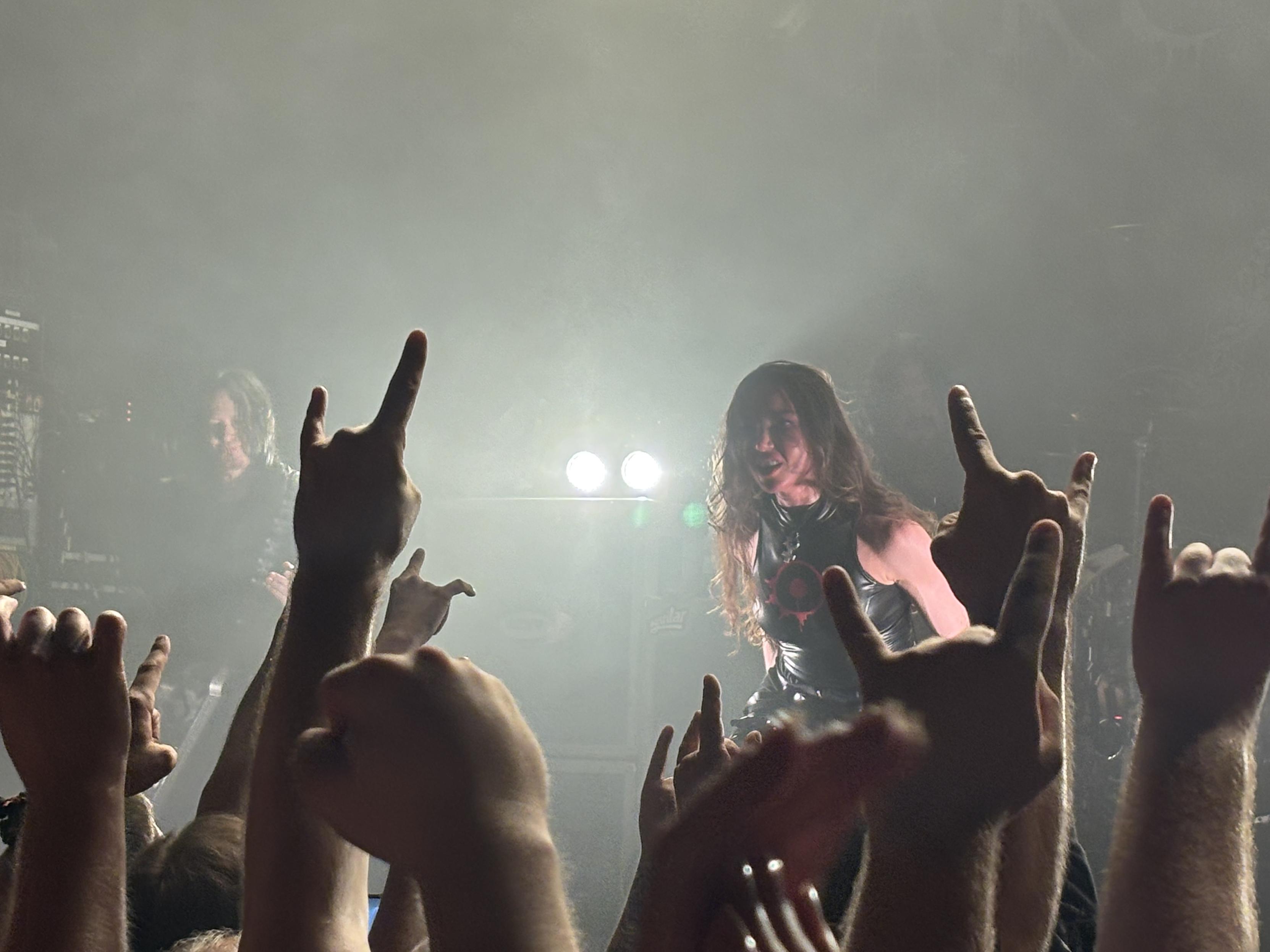 En bild från en metalkonsert. I förgrund flera höjda händer som gör horntecknet. Bortom det en kvinnlig sångare [Lauren Hart] och i bakgrunden en manlig gitarrist [Michael Amott].