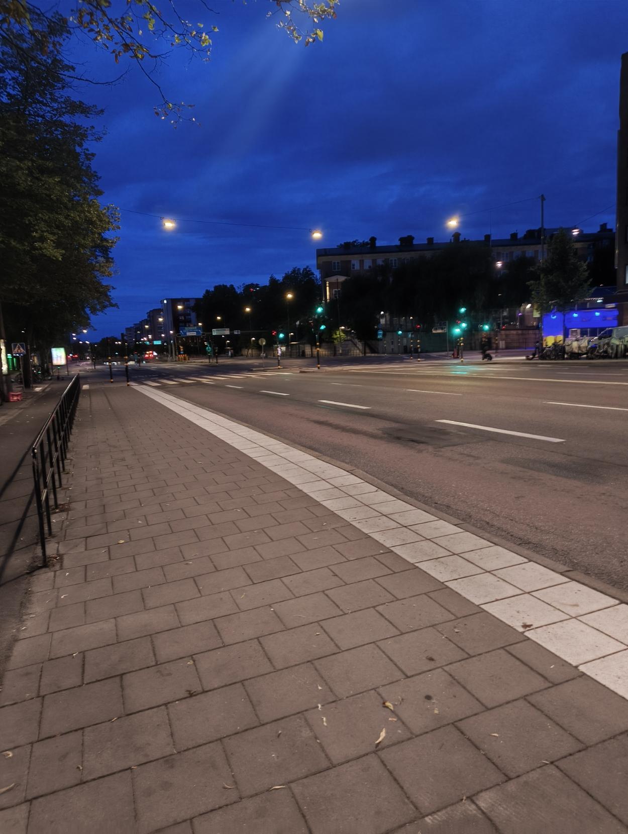 An empty city street at twilight under a deep blue sky, featuring a paved sidewalk along a wide, multi-lane road illuminated by glowing streetlights and traffic signals.