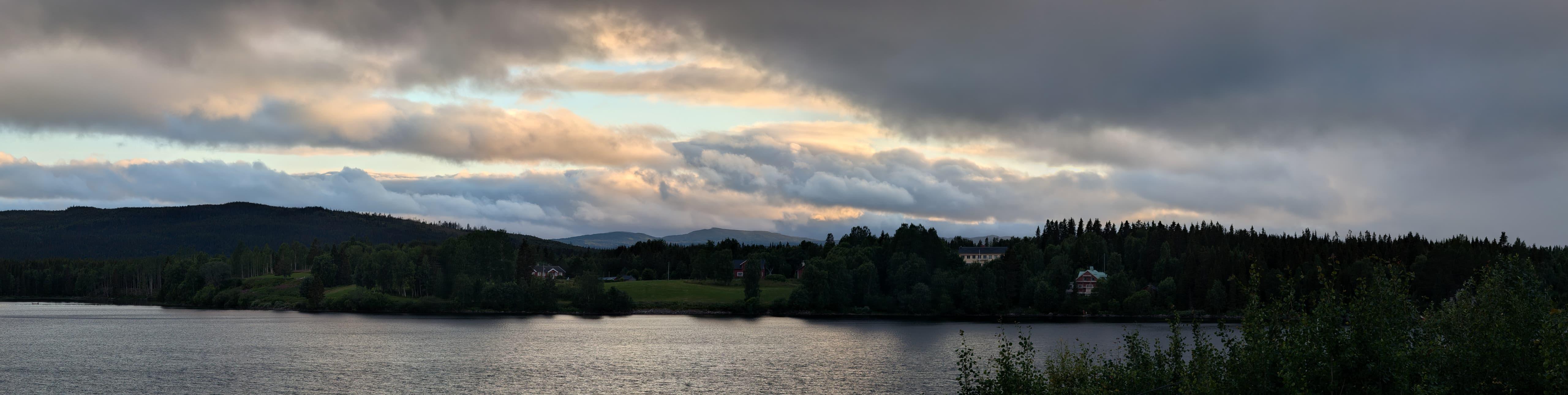 A panorama of the sun setting over a mountain lake.

In the right foreground there are a few trees. The lake is calm. On the other side of the lake are trees and a few houses. On the distance a couple of mountain tops can be seen.

The clouds are hanging low and creates a beautiful contrast between orange of the setting sun and the grey/blue of the water and clouds.