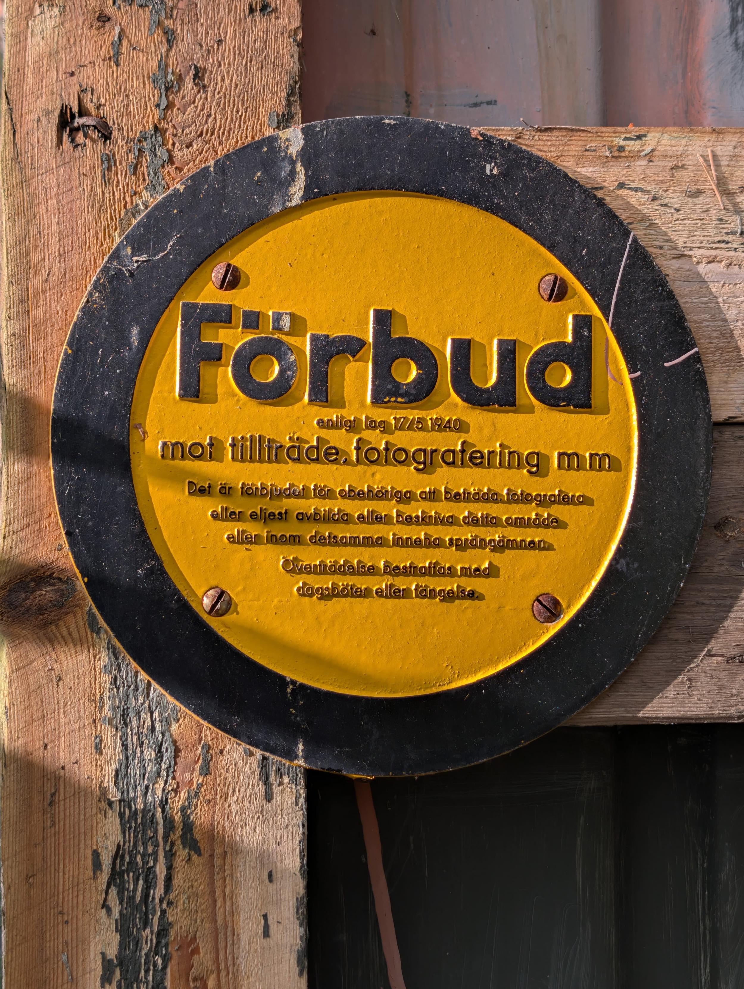 Foto av en mycket gammal skylt. Rund, gul med svart kant och svart text. Texten lyder:

"Förbud
enligt lag 17/5 1940
mot tillträde, fotografering mm

Det är förbjudet för obehöriga att beträda, fotografera eller eljest avbilda eller beskriva detta område eller inom detsamma inneha sprängämnen.

Överträdelse bestraffas med dagsböter eller fängelse."