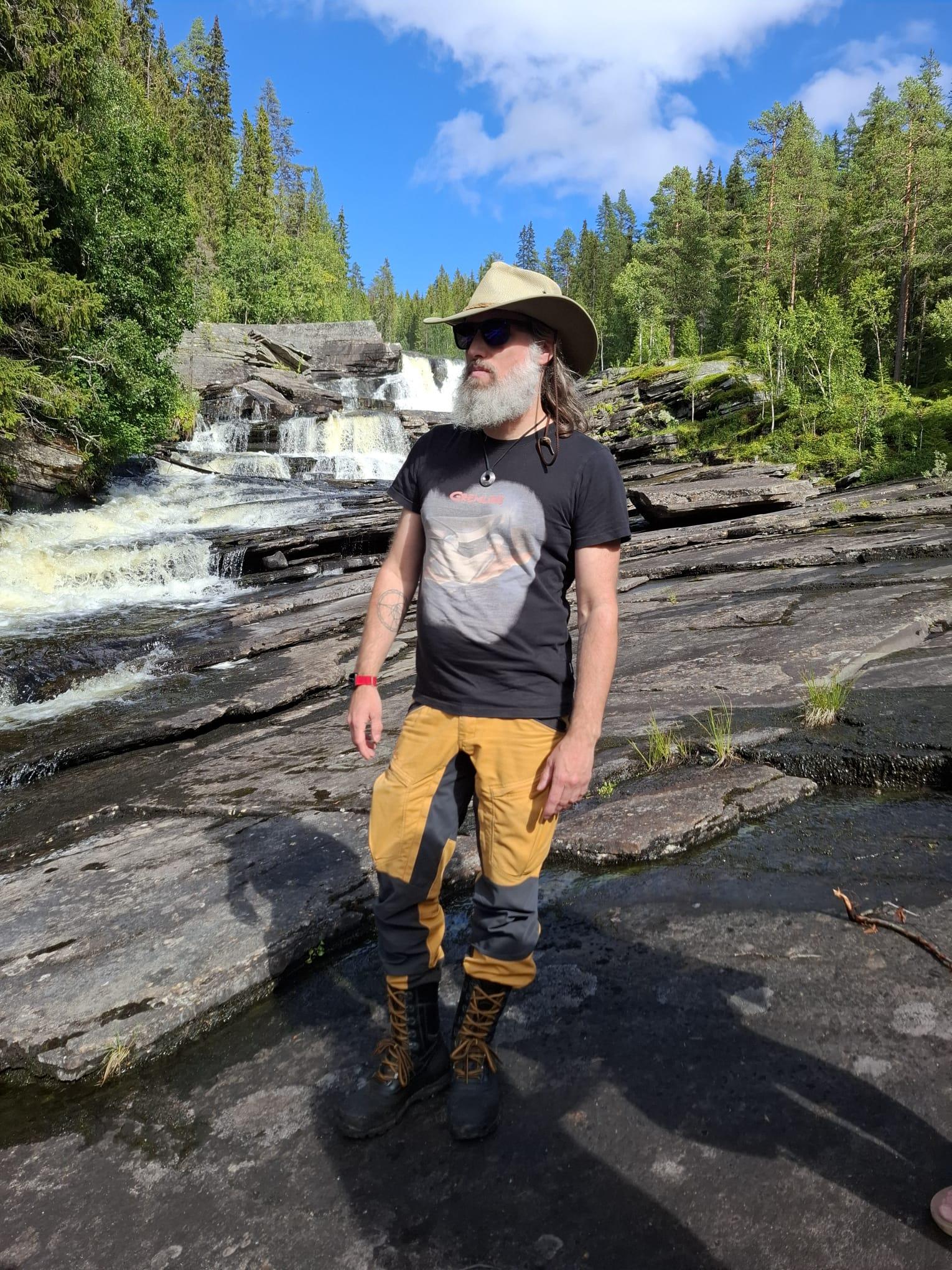 A photo of a man standing on large flat rocks next to a flowing rapid.

Behind him a series of waterfalls can be seen, surrounded by spruce and birch trees. The sky is blue with a few tufts of white clouds.

The man is dressed in hiking boots, dark yellow wilderness pants with black parts over knees and inside of the thighs, a black t-shirt with a Gremlins print (Gizmo sticking his small paws out from a box carried by two hands, the movie poster I think) and a light coloured wilderness style hat with a wide brim.

The man is wearing sunglasses, has a large and bushy white beard and shoulder length hair currently held back by the hat. A tattoo is visible on the inside of the right forearm and if you look closely there's one on the left as well.

He's looking at something outside the image, to the left.