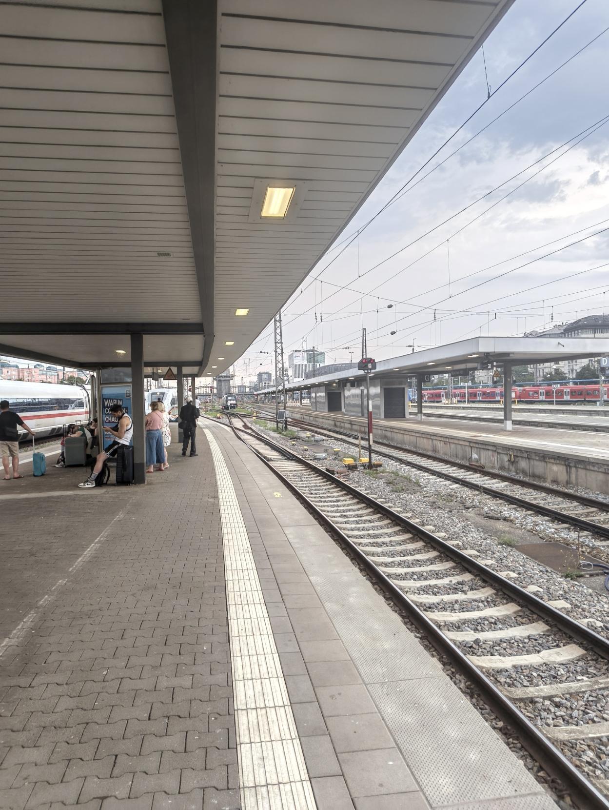 An outdoor train station platform under an overhead canopy, with passengers waiting alongside luggage, train tracks running down the middle, and a white ICE train stopped on the left.