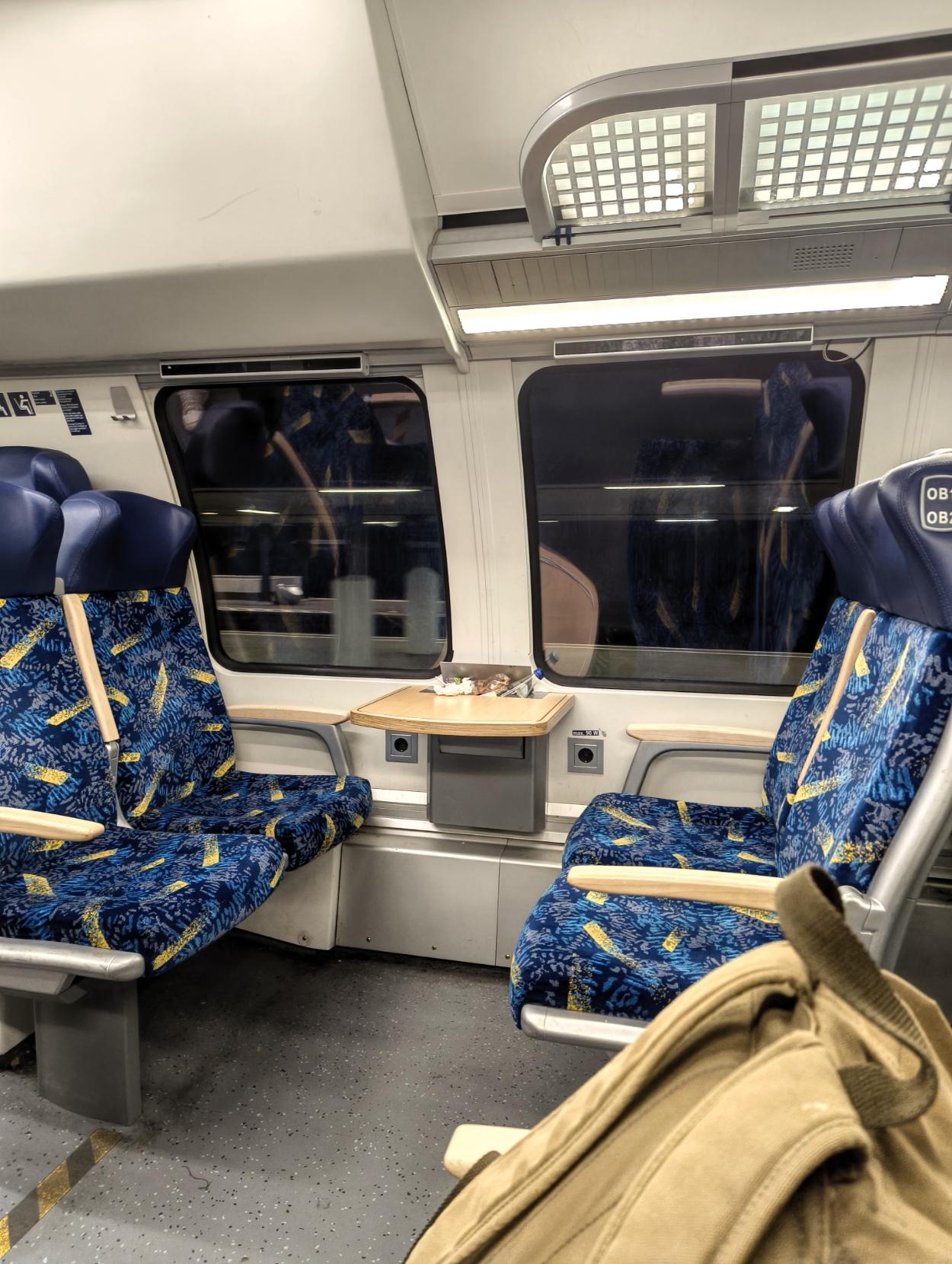 ​The interior of a train compartment featuring four facing blue-patterned fabric seats, a small wooden table between them by the window, and a canvas backpack in the foreground.