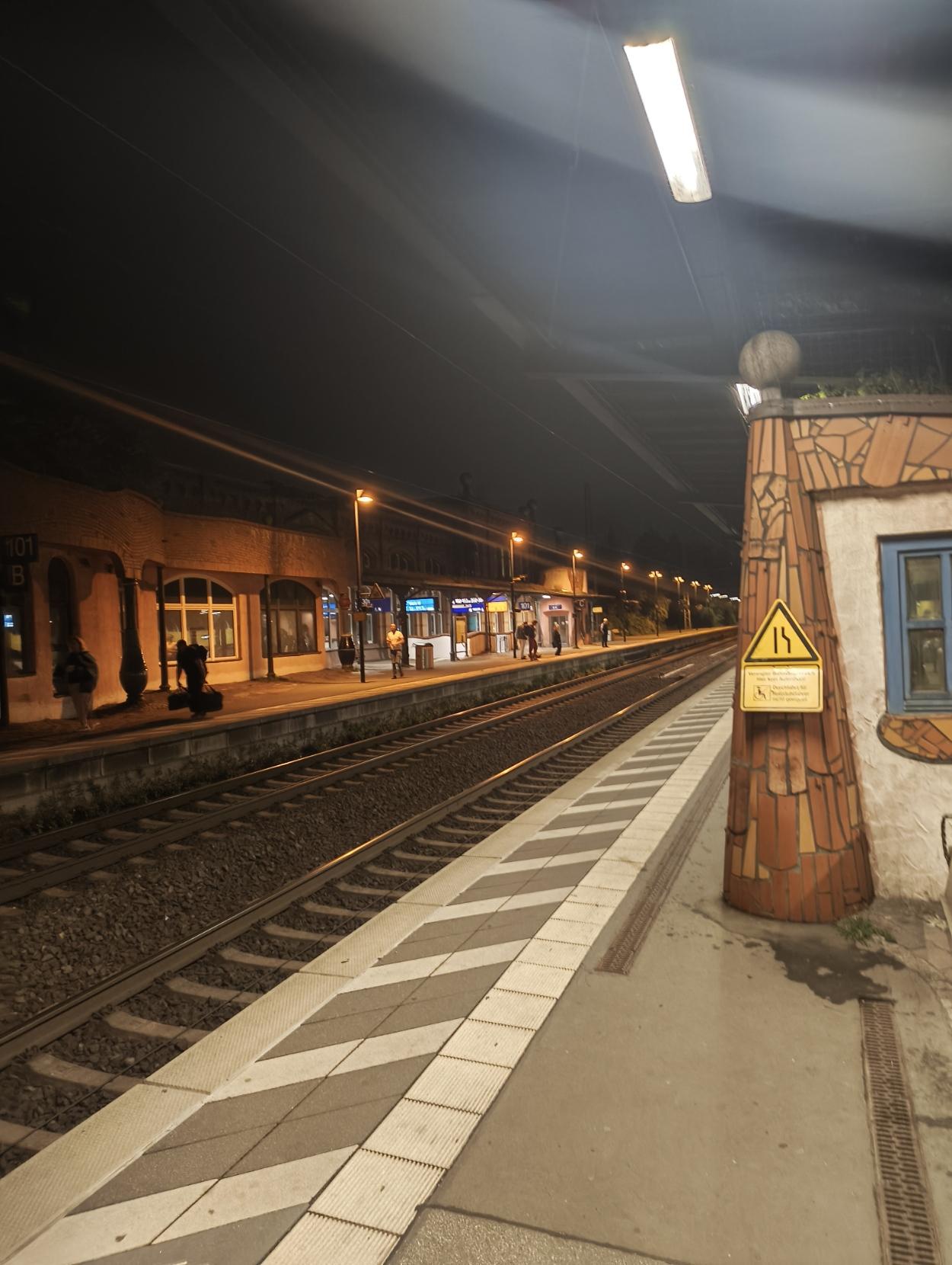 Nighttime view of an outdoor train station platform with train tracks, glowing streetlights, waiting passengers, and a distinctive mosaic-tiled column in the foreground.